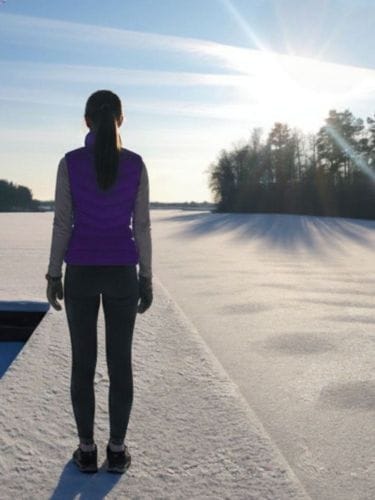 Przystanek Wellness - kobieta stojąca na oszronionym pomoście, przed nią zamarznięte jezioro, w tle drzewa, słoneczny, zimowy dzień